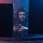 A mysterious hacker in a dark room using a smartphone between two monitors