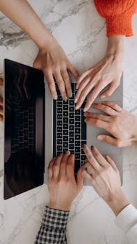 A top-down view of multiple hands typing on a laptop keyboard on a marble desk, indicating teamwork.