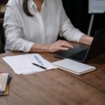 Businesswoman in white shirt working on laptop with papers at wooden desk.