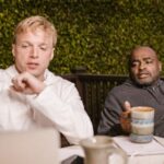Two adults collaborating outdoors with coffee mugs, discussing work on a laptop.