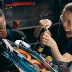 Two students working together on building a robot in an industrial setting, wearing protective goggles.