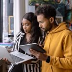 Two young adults collaborating using laptop and tablet indoors, showcasing teamwork and technology.