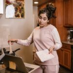 Woman multitasking in her kitchen with a laptop and notebook, cooking and working from home.