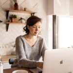 Young female in gray sweater sitting at wooden desk with laptop and bottle of milk near white bowl while browsing internet on laptop during free time at home