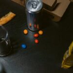 A casual setup with pizza slice, soda can, candy and open snack bag on dark table.