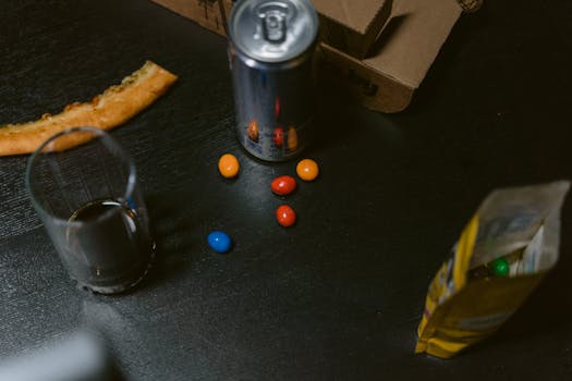 A casual setup with pizza slice, soda can, candy and open snack bag on dark table.