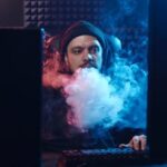 A man sits at a desk, vaping, while focused on computer screens in a dimly lit room.