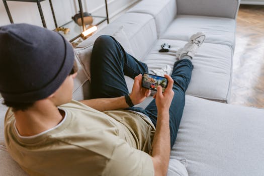 A young man in casual wear playing a mobile game while relaxing on a sofa indoors.