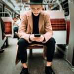 Full body of stylish concentrated young male in trendy hat and coat sitting on passenger seat in train and messaging on mobile phone