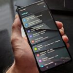 Hand holding smartphone displaying settings screen with various options evident.