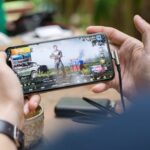 Person playing PUBG on smartphone outdoors with charging cable attached.