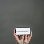 Symbolic image of addiction showing hands in chains holding a phone displaying 'Gambling Addict'.