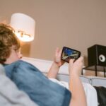 Teen lying on sofa playing video games on smartphone indoors, surrounded by modern décor.