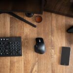 Top view of black gamepad with mouse and keyboard on wooden desk with monitor in apartment in daytime