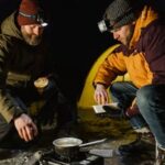 Two men cooking at a campsite on a frozen lake during a winter night.